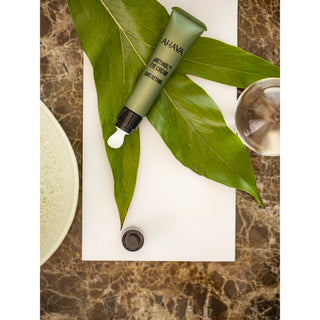 A tube of Ajava Antioxidant & Eye Cream lies diagonally on a white rectangular board, surrounded by large green leaves, with a small open cap below and a marble-patterned surface around it.