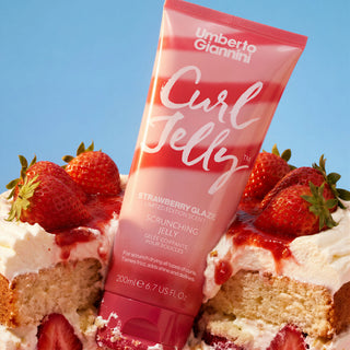 A tube of Umberto Giannini Curl Jelly Scrunching Jelly in Strawberry Glaze flavor, styled on top of a strawberry and cream cake against a solid blue sky background.
