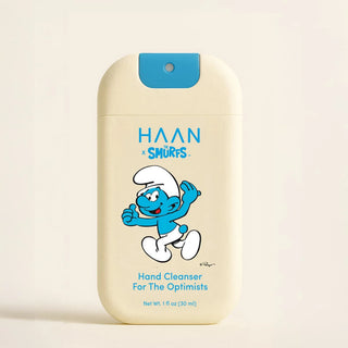 A 1 fl oz (30 ml) bottle of HAAN x Smurfs Hand Cleanser. The bottle is cream-colored with a blue cap and features a cartoon illustration of Smurfy smiling.