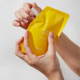 A person's hands holding a bright yellow NAAN Citrus Noon refill pouch with a white cap, shown against a clean light gray background.