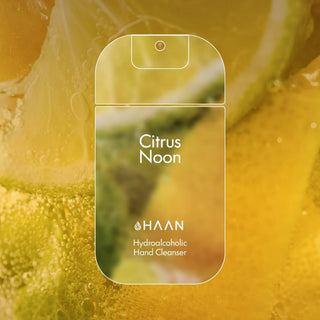 A bottle of HAAN Citrus Noon hydroalcoholic hand cleanser, designed to look like a smartphone outline, set against a vibrant yellow and green citrus background with visible bubbles.
