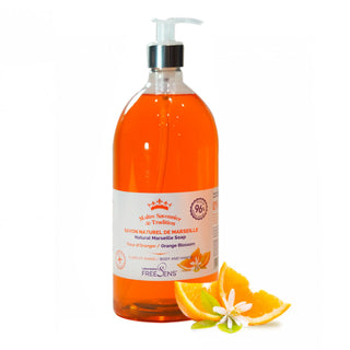 A bottle of Free Lens Marseille Soap body wash in orange blossom scent, filled with orange-colored liquid and topped with a white pump dispenser. It is displayed on a clean white background with orange slices and a blossom garnish at the base.