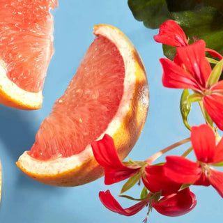 A close-up still life of a pink grapefruit slice and another grapefruit wedge beside bright red flowers on a light blue background.