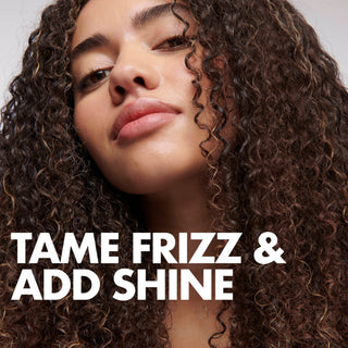 Close-up of a woman with voluminous dark brown curly hair and light golden highlights, with large white text reading 'TAME FRIZZ & ADD SHINE' across the lower portion.