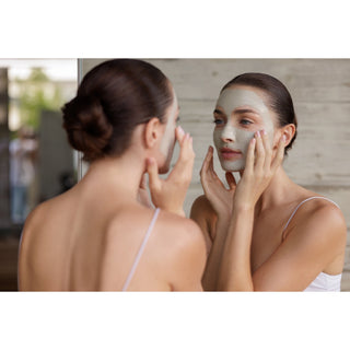 A woman applies a light green facial mask while looking at her reflection in a mirror, wearing a white spaghetti-strap tank top against a light wood-paneled background.