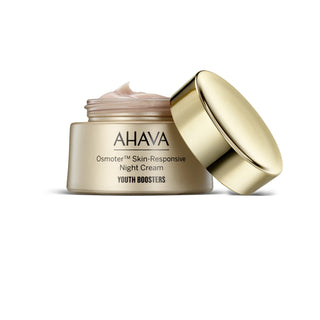 AHAVA Youth Boosters Osmoter Skin-Responsive Night Cream in a metallic gold jar with its lid leaning against it, showing a pale pink cream inside, on a white background.