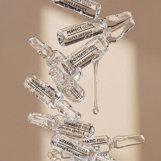 Several clear glass ampoules labeled PERFECT GLOW Super Ampoule, Enzymatic Peel Super Ampoule, and Vitamin C Ester Super Ampoule are arranged diagonally against a warm beige background, with one ampoule showing a liquid drop dripping from the top.