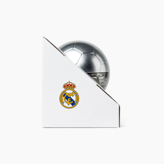 A silver metallic soccer ball trophy partially hidden behind a white triangular display stand featuring the Real Madrid crest, on a clean white background.