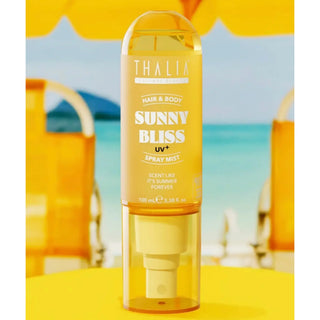 A bottle of Thalia Sunnys Bliss UV+ Spray Mist hair and body product, shown in a translucent yellow bottle with a clear cap, set against a blurred beach background with yellow umbrellas.