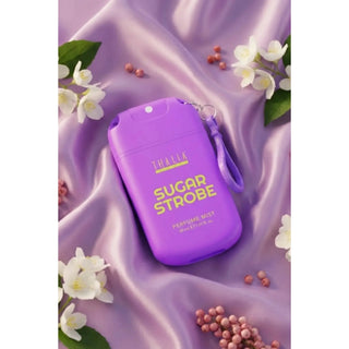 A purple THALIA Sugar Strobe perfume mist bottle with a wrist strap, styled on a draped lavender satin fabric with white flowers and small pink berries.