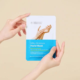 A person holds a flat sheet mask package labeled 'Milky Moisture Hand Mask' against a plain beige background. The package is white on top with a photo of hands, and blue on the bottom with product text.