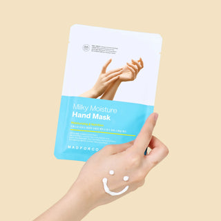 A hand holds a stand-up pouch of Madforcen Milky Moisture Hand Mask against a solid beige background. The packaging is white and light blue, featuring an image of hands, and a small amount of white cream is smeared on the back of the holding hand.
