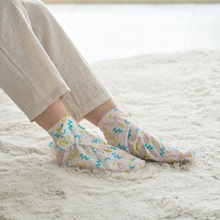 A person wearing pastel multicolor abstract scribble socks with cream-colored textured pants, sitting on a fluffy white rug.