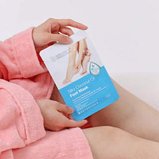 A person in a pink terry robe holds a sheet mask package labeled 'Silya Coconut Oil Foot Mask' against a clean white background.