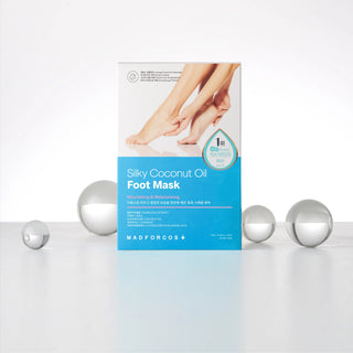 Mad For Cos Silky Coconut Oil Foot Mask packaging box standing upright on a white surface, with several reflective silver spheres arranged around it against a clean white background.