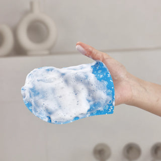 A hand wearing a blue silicone cleaning glove covered in white foam, shown against a blurred bathroom background.