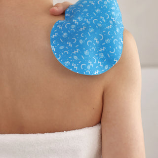 A close-up of a person's upper back and shoulder with a blue cooling pad featuring a white abstract pattern applied to the shoulder. A white towel is wrapped around the lower back.