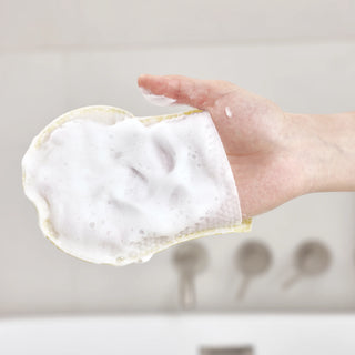 A hand holding a yellow sponge covered in thick white foam, with a blurred bathroom background showing faucets.