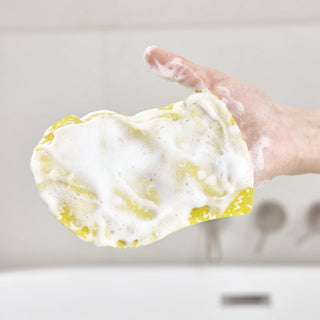 A hand holding a yellow sponge covered in thick white foam, with a blurred bathroom sink and faucet in the background.