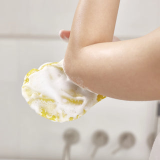A close-up of a person's forearm and hand holding a yellow sponge covered in white foam, with a softly blurred bathroom background.