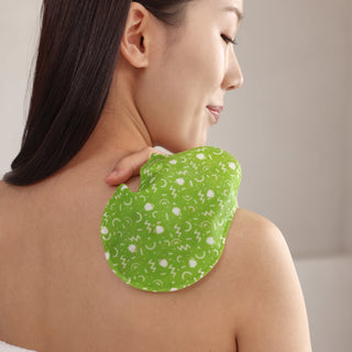 A woman shown from the back with her neck and shoulder exposed, holding a green shoulder massage or heat pad with a white abstract pattern against her upper shoulder.