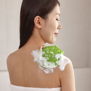 A woman with her back turned applies a green, bubble-covered shoulder scrub to her upper shoulder and neck. She is wrapped in a white towel, set against a soft, neutral-toned bathroom background.