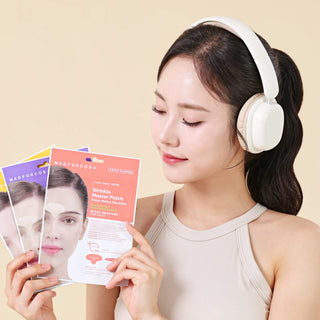 A woman wearing white over-ear headphones holds three colorful skincare product packets in front of her face against a soft beige background. The packets appear to be Madforcosa Wrinkle Wrangler products.