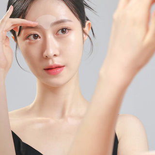 A close-up of a woman with a clear, semi-translucent circular eye patch applied to her forehead, wearing a black top against a solid light gray background.