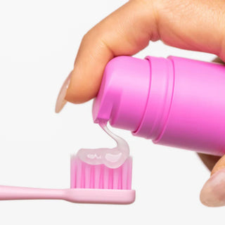 A close-up of a hand squeezing bright pink toothpaste from a matching pink tube onto a pink toothbrush against a clean white background.