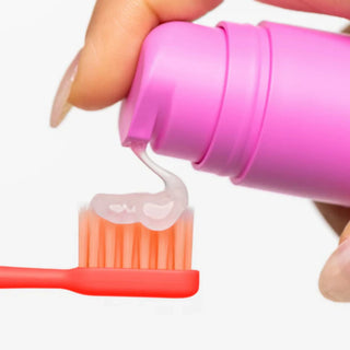 A close-up of a hand squeezing a bright pink tube to dispense clear toothpaste onto a coral-red toothbrush against a plain white background.