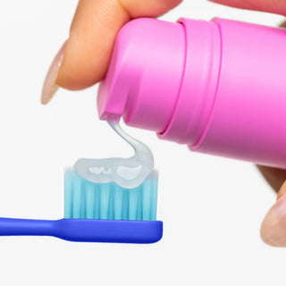 A close-up of a hand squeezing a bright pink pump bottle to dispense clear toothpaste onto a blue toothbrush against a clean white background.