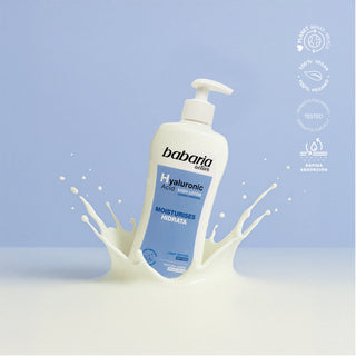 A bottle of babaria Golds Hyaluronic Acid moisturizer stands upright in a milk splash against a soft light blue background. White circular certification-style icons appear on the right side.