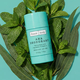 A turquoise deodorant stick labeled 'frank body AHA DEODORANT' placed on a bed of green mint leaves and eucalyptus against a turquoise background.