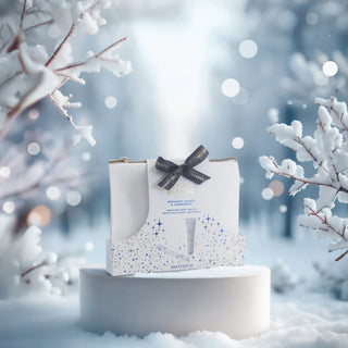 A boxed bath product displayed on a round pedestal in a snowy outdoor scene. The package is white with blue and gold speckled accents, tied with a dark ribbon, and the visible text includes 'BATHING' and 'PREMIER COLD REMOVAL'.