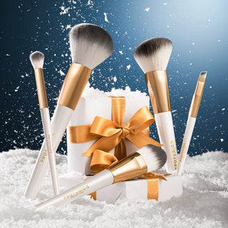 A set of five PA:VISION professional makeup brushes arranged around a white gift box with a gold ribbon and bow, set in white snow against a dark blue background with falling snow.