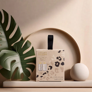 A boxed bath gift set labeled 'Luxury Bathing' stands on a beige platform with a large green monstera leaf on the left and a round white object on the right. The box has a beige leopard print pattern with a black ribbon handle and small product images on the front.