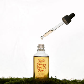 A clear glass dropper bottle labeled 'Colagen Density Drops' by Umberto Giannini, with a glass pipette above it dispensing liquid, set on green moss against a white background.