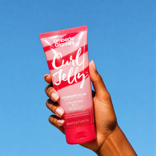 A hand holding a tube of Umberto Giannini Curl Jelly Strawberry Glaze hair scrunching jelly against a solid blue sky background.