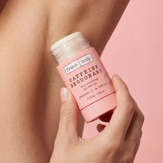 A hand holding a pink Frank Body Caffeine Deodorant stick against a model's armpit and a solid pink background.