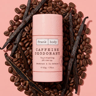 A pink Frank Body caffeine deodorant stick surrounded by coffee beans and vanilla beans on a pink background.
