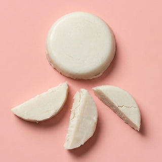 A round off-white solid bar, likely soap or wax, shown whole with three cut wedges beside it on a soft pink background.