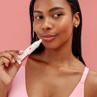 A woman with dark hair wearing a pink ribbed top holds a white cosmetic tube with red accents near her lips, set against a solid pink background.