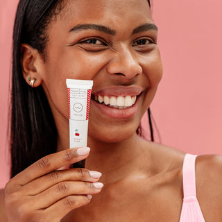 A smiling woman holds a white tube of Fresh Lip Balm against a solid pink background. The tube is white with red text and a small red cherry graphic, and the brand name Fresh is visible.