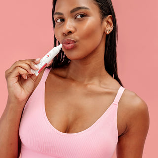 A woman wearing a pink ribbed tank top applies a white lip balm with red text to her lips, set against a solid pink background.