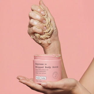A pink tub of Fruit Body Express-o Whipped Body Scrub held by two hands, with one hand showing the thick beige scrub texture against a solid pink background.