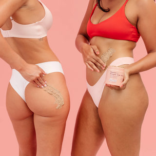 Two models in pastel pink and coral bikinis demonstrate the application of a beige body scrub on their hips, with one holding a pink jar of 'Nourishing & Nourishing Body Scrub' against a solid pink background.