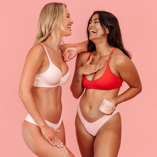 Two women posing in matching light pink and red bikini sets against a solid pink background, with one holding a jar of exfoliating scrub and sand visible on their skin.