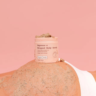 A pink jar of Express-o Whipped Body Scrub sits on top of a person's bare back, which is covered in the scrub. The background is a solid pink color.