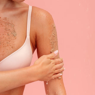 A close-up of a woman's upper torso and arm wearing a white bra, with a beige exfoliating scrub or sand applied to her skin and being rubbed onto her arm against a soft pink background.