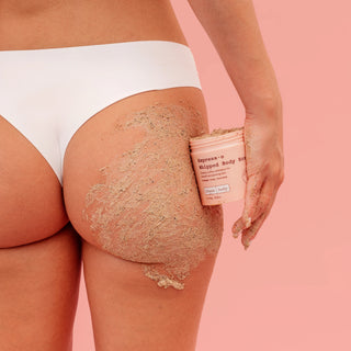A close-up of a person wearing white underwear and applying or showing a beige textured body scrub on the buttocks, holding a pink jar labeled 'Impress-o Whipped Body Scrub' against a solid pink background.